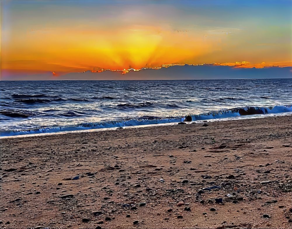  A sunset on the beach with orange rays coming out from behind the clouds close to the horizon The clouds are outlined by the bright yellow sunlight as the wave come crashing on the beach sand that is almost covered with seashells. 