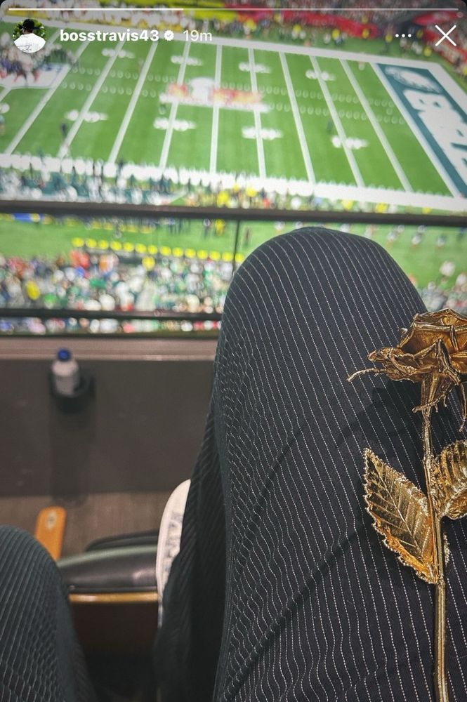 a screenshot of ross travis instagram story which is a picture of his POV at the super bowl. In the bottom half it shows his thigh and he’s wearing black pants with white pinstripes and a gold rose is sitting on his leg