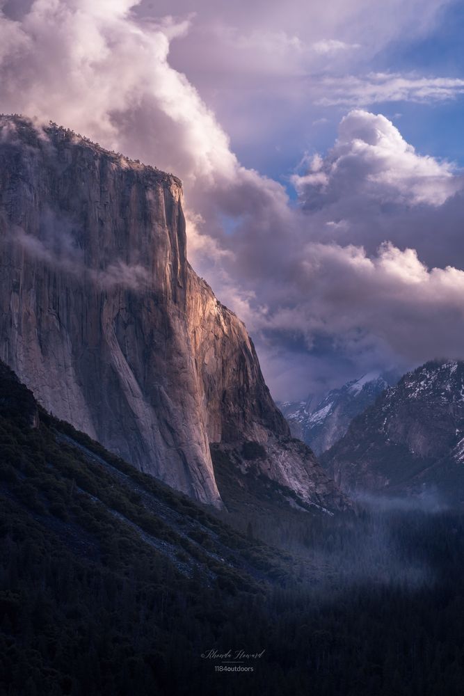 Early morning light catches the face of a large rock formation known as El Capitan. Below is a valley of mostly evergreen trees and some fog along the Merced River. The sky has dramatic cloulds with some blue sky breaking through. In the distance some snow can be seen on the ground.