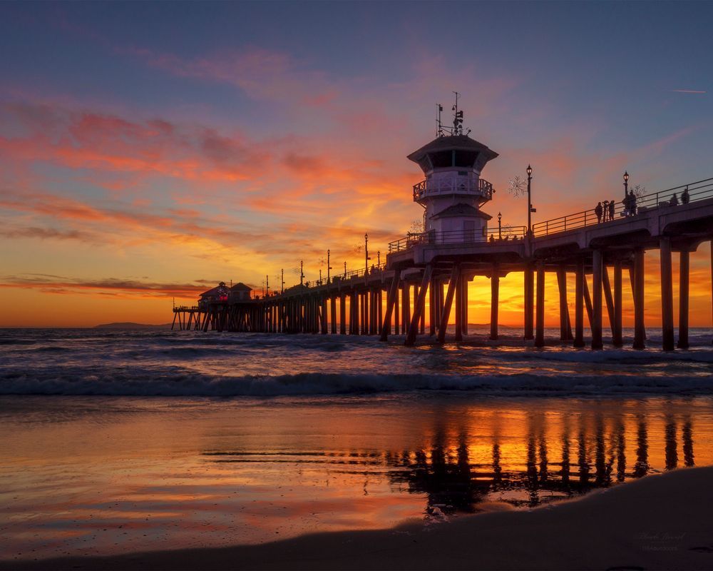 View of a pier silhouetted against a sunset with golden, orange, pink, and purple hues. There's a partial reflection of the pier on the water.