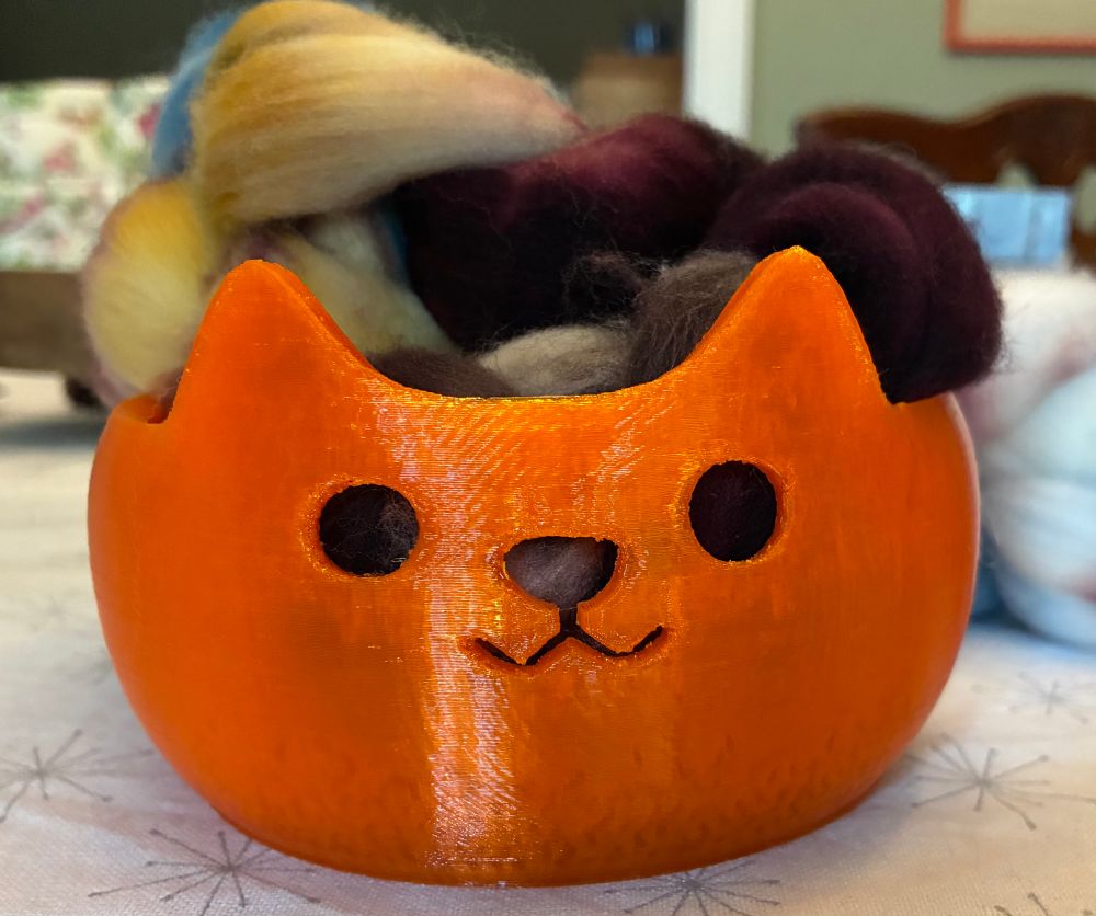 Cat head shaped yarn bowl from The Foldout Cat. The yarn bowl is bright pumpkin orange with cut out holes for the cat’s eyes, nose, & mouth. 