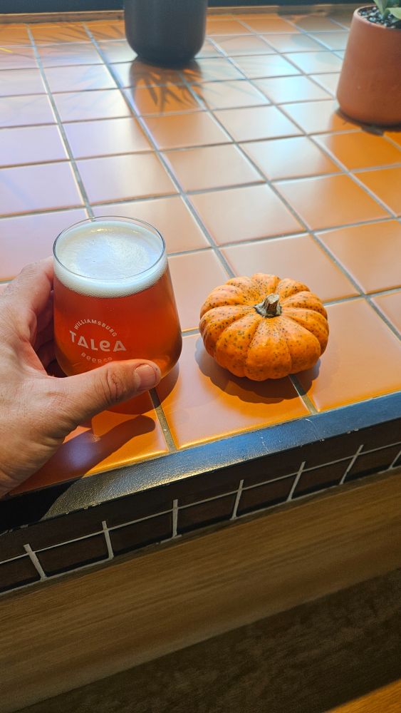 A man holds a dark colored beer alongside a mini pumpkin on a tiled surface