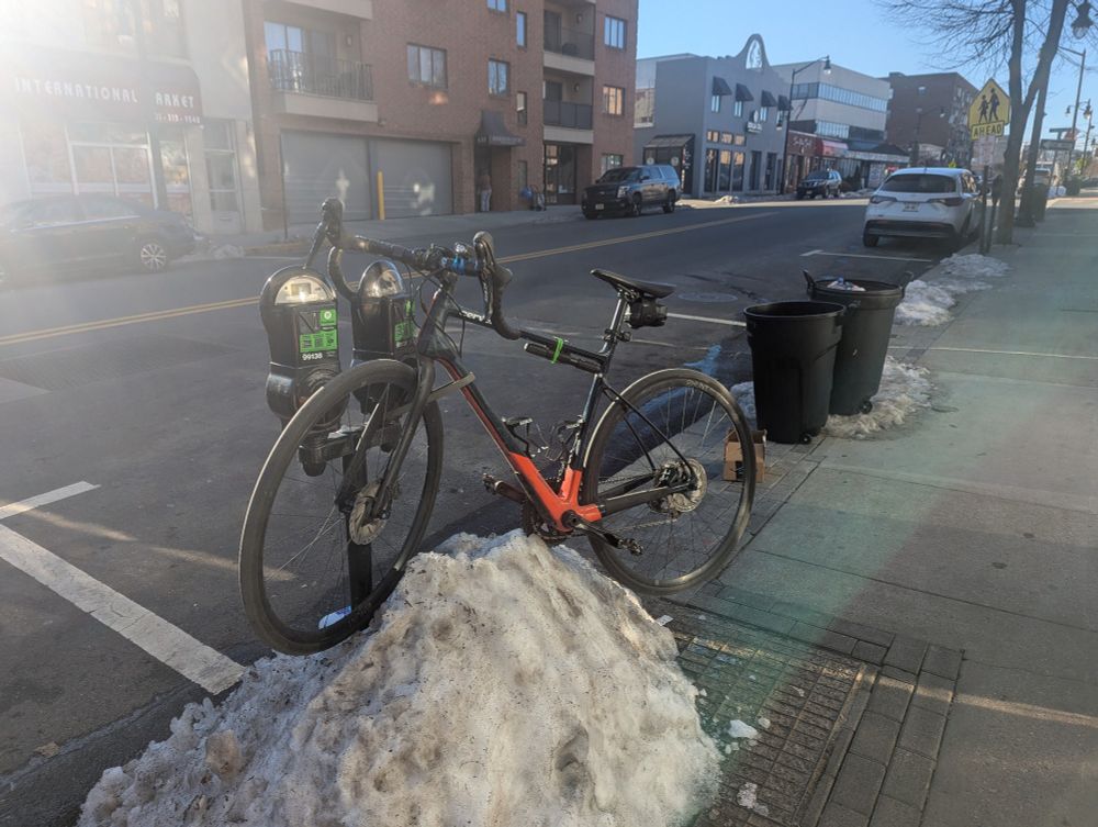 A road bike locked to a tandem parking meter with a meter-high pile of snow and ice at the base of it, bike perched on the snow