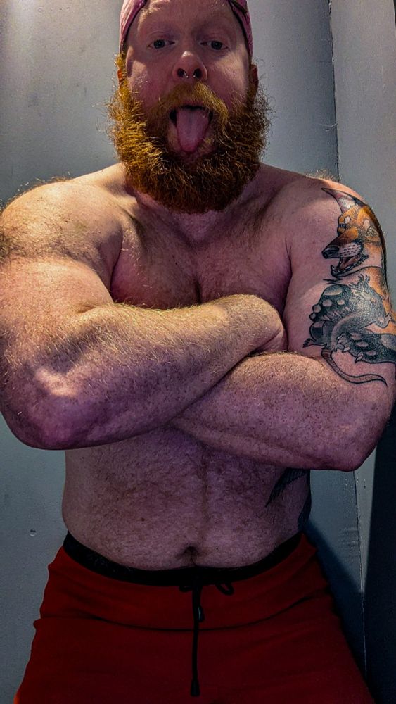A topless man with a thick ginger beard flexes in front of the camera, crossing his arms over his chest while sticking his tongue out.
