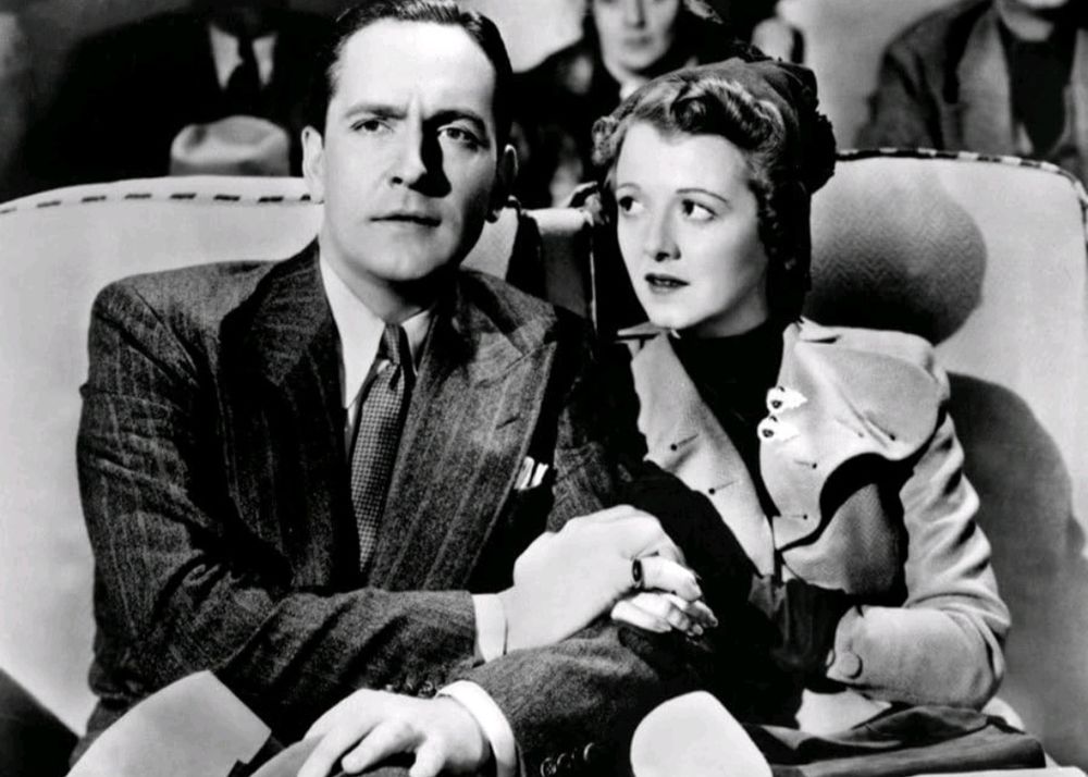 Fredric March & Janet Gaynor, sitting with arms linked.