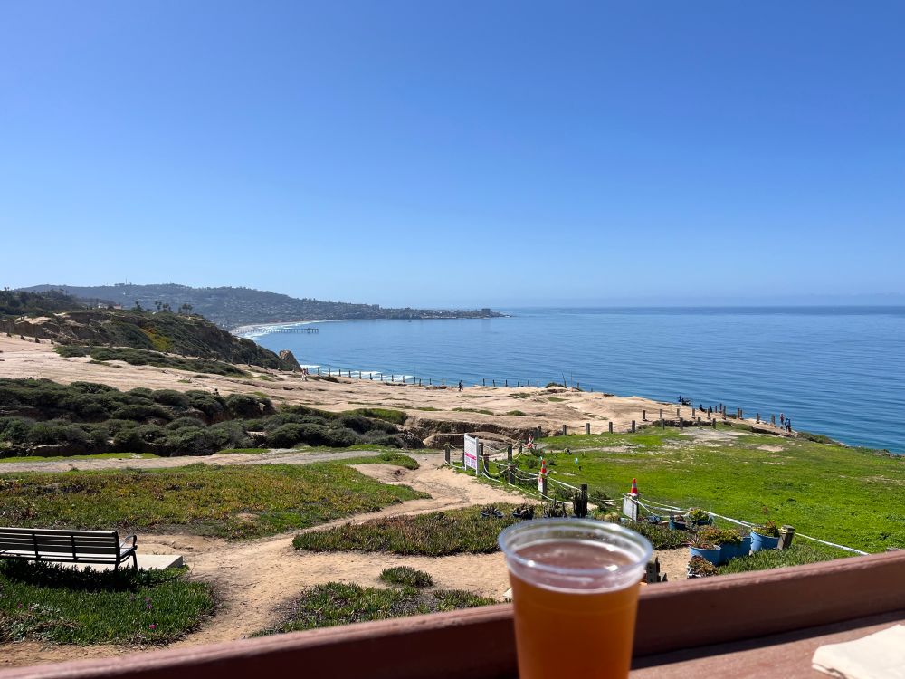 A picture of an ocean landscape:  In the foreground is a beer sitting on a bar rail.  Behind the rail is a grass and dirt field that descends towards the Pacific Ocean.  In the background is a view of La Jolla.