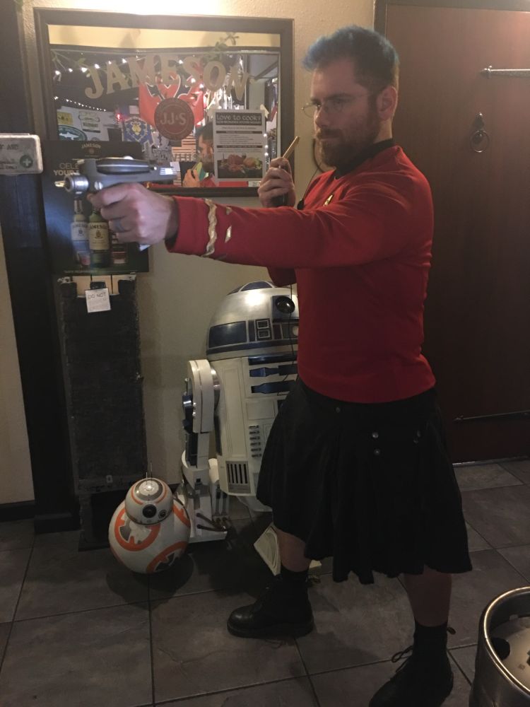 Guy in a black kilt and TOS Engineer tunic pointing a phaser in front of some droids
