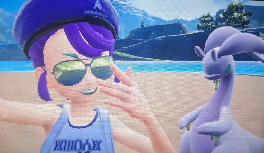 A photo from a Nintendo switch of a beach scene with a person with purple hair and aviator shades taking a selfie with a gray and purple Pokemon, goodra. The goodra is asleep while the person is smiling at the camera 