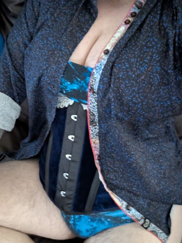 A white person with dark facial and body hair wearing an underbust corset with blue velvet and embroidered white wings and a blue floral dress button down with blue galaxy underpants. 