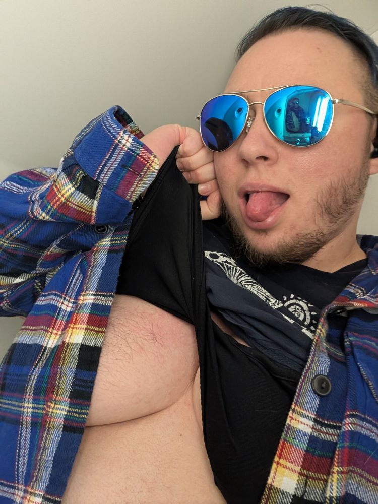 A white person in sunglasses with dark body and facial hair lifting their button down flannel and dark blue tee shirt to show their under and side boob. 