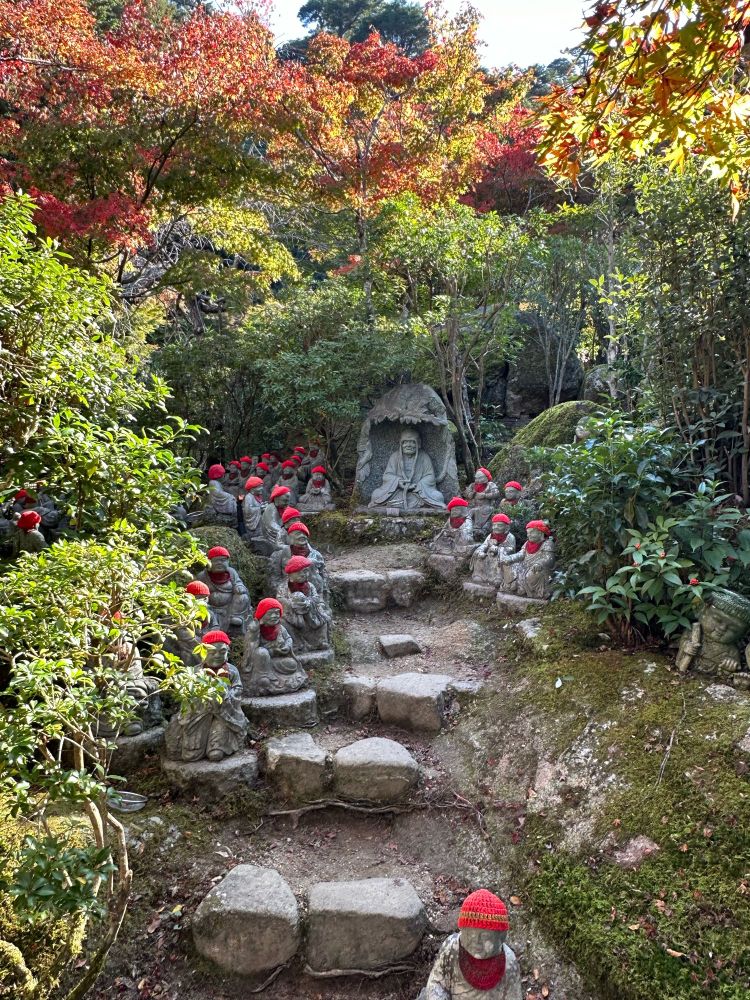 A path through a garden in Daisho-in temple. The sides of the path are lined with Rakan, tiny statues of Buddhist monks. Each of them is wearing a red knit cap and some have little red scarves as well, offerings to keep them warm. In the center at the end of the path a larger more intimidating statue sits cross legged in a relief. He has long eyebrows that stretch down to his legs.
