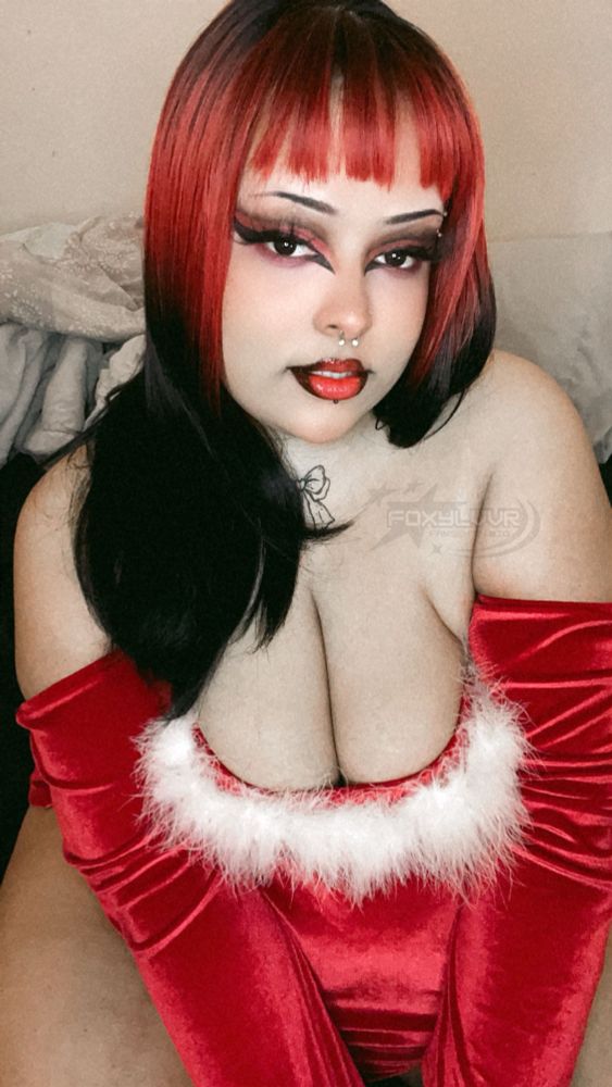 a picture of an ftm fem presenting fat person who’s Black, wearing a red santa dress with white fluff at the top hem of the collar and red sleeps that are off the shoulder. his arms are crossed infront of him pushing his boobs together while you can see his bare hips from behind his arms in frame. he has long red and black hair down his shoulders, and red and black makeup on his eyes with red lipstick. he’s looking into the camera seductively, with his head tilt to the side