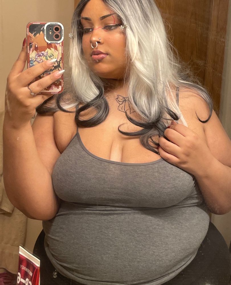 theo, an ftm fem presenting fat person who’s Black, wearing a grey tank top and standing in front of a mirror. he’s holding his hair, it’s white and black waves down his shoulders 
