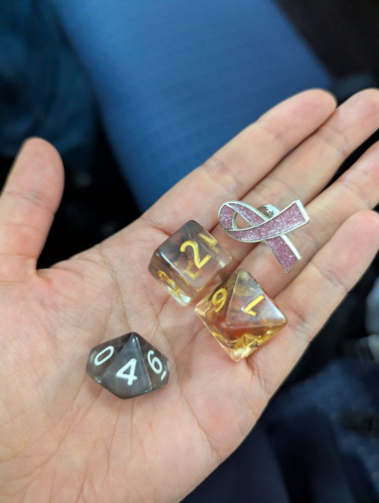 A few dnd dice and a pink breast cancer pin laying on an outstretched hand