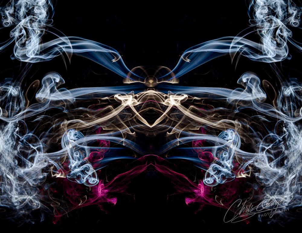 A symmetrical abstract pattern created from swirling smoke in white, blue, tan, and pink tones against a black background. The image shows curling tendrils and layered wisps of smoke forming mirrored shapes that resemble wings or flowing energy. Fine details of the smoke lines appear crisp and textured.