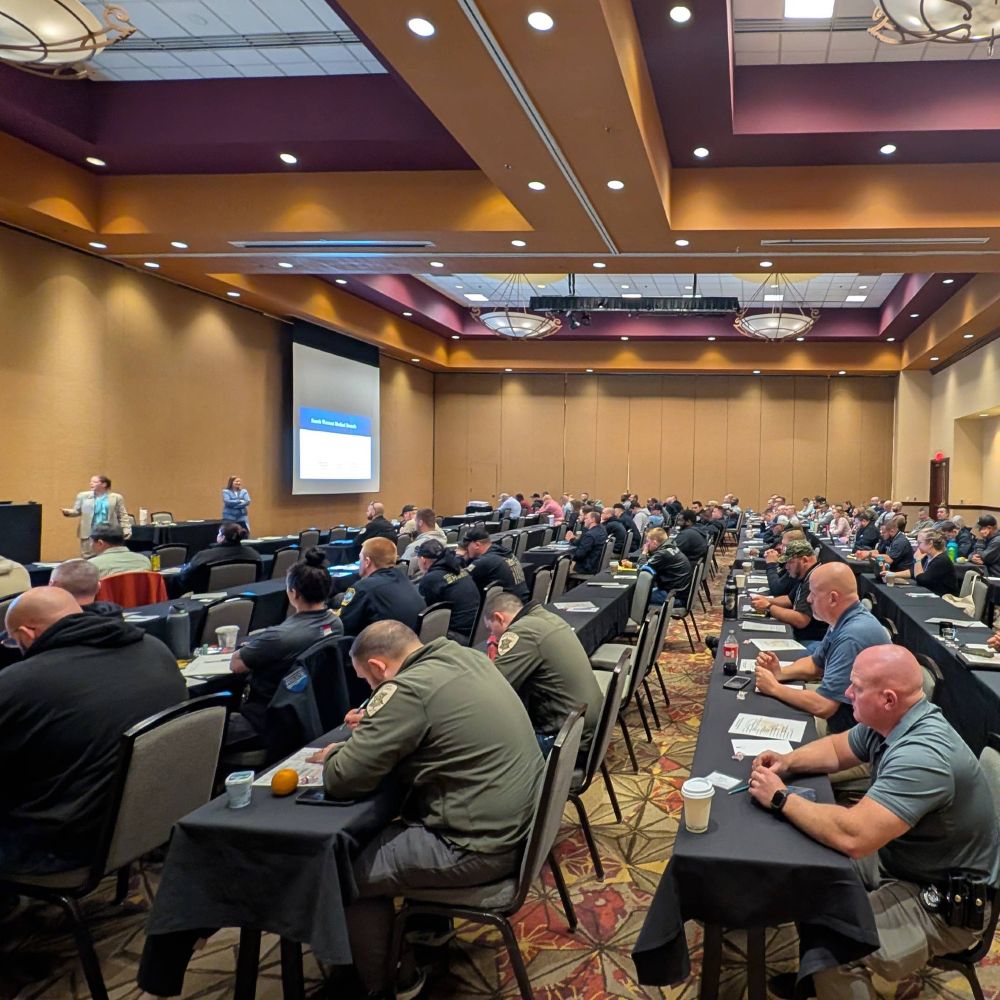 A large group of people are seated at tables in a conference room, facing a speaker at the front. Attendees are dressed in a variety of clothing, and some are taking notes or looking at papers. The room is well-lit with warm-toned walls and patterned carpeting.