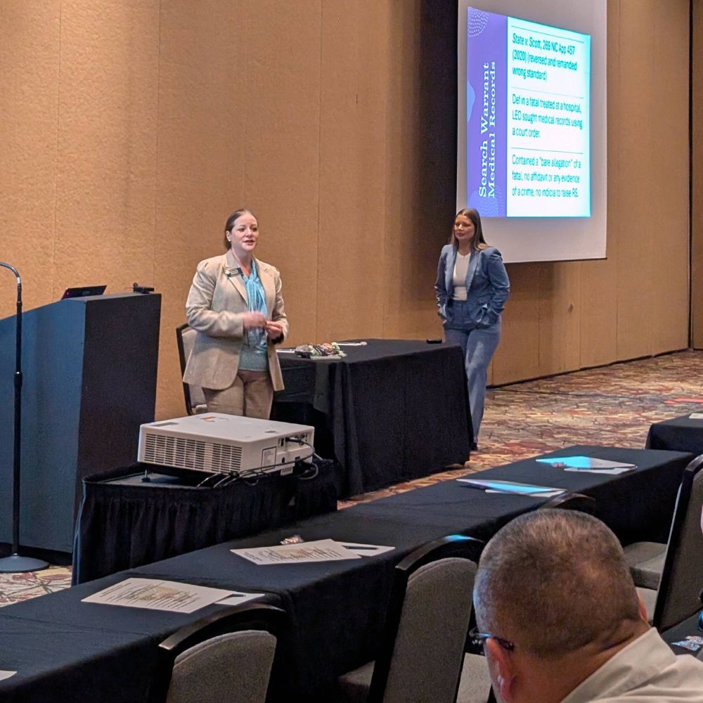 Two people presenting in a room with a projector screen. The person on the left is speaking and has light-toned skin and light-colored hair. The person on the right has light-toned skin and medium-length brown hair. The back of a person's head is visible in the foreground.