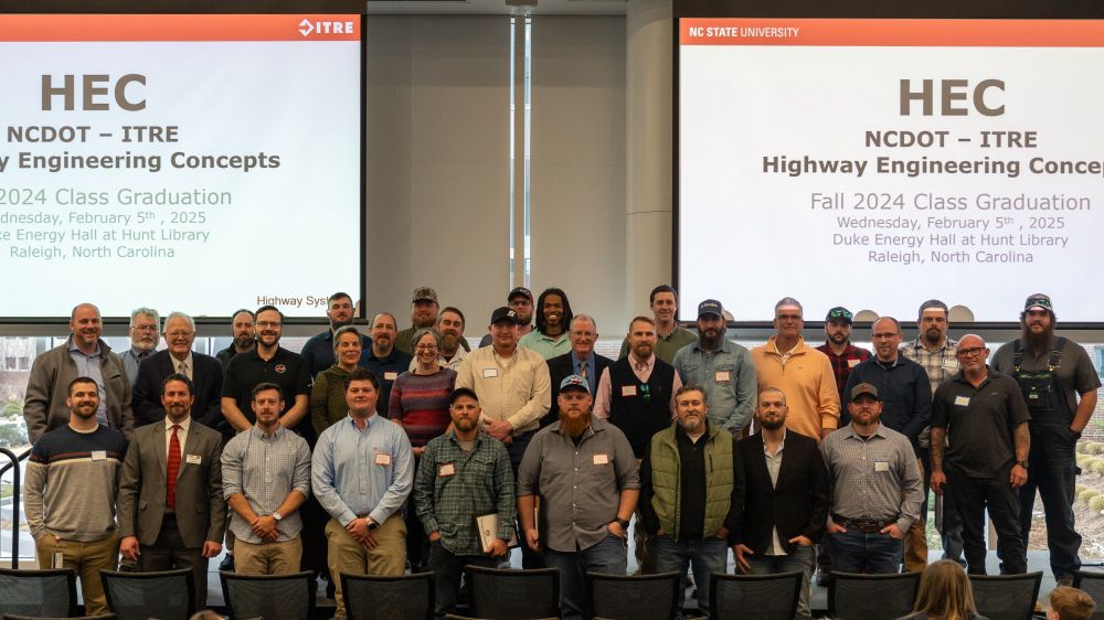 About 30 people posing for a group photo at the front of a large room. Projector screens behind them read "HEC. NCDOT - ITRE. Highway Engineering Concepts. Fall 2024 Class Graduation. Wednesday, February 5th, 2025. Duke Energy Hall at Hunt Library. Raleigh, North Carolina."