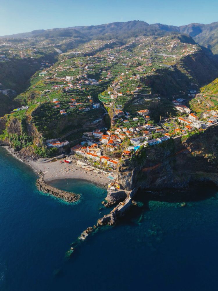 Drone shot from coastline of Madeira, seeing a small town and a beach with mountains in the background. Sunny clear sky