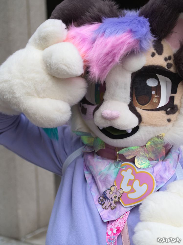 A brown cheetah fursuiter looking towards the viewer, raising one hand to lift their purple and pink hair. The hand partially covers their rainbow eye, leaving their brown eye clear