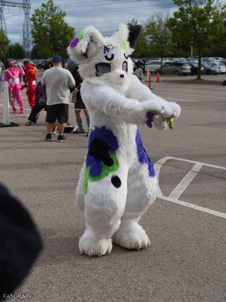 White canine (dog?) fursuiter with floppy ears and green and blueberry ink splatter patterns on the waist and thihgs, dancing in a parking lot section cleared of cars. Their eyes are plastic domes that have fogged up. There are several cosplayers congregating in the background, and parked cars further in the distance. 