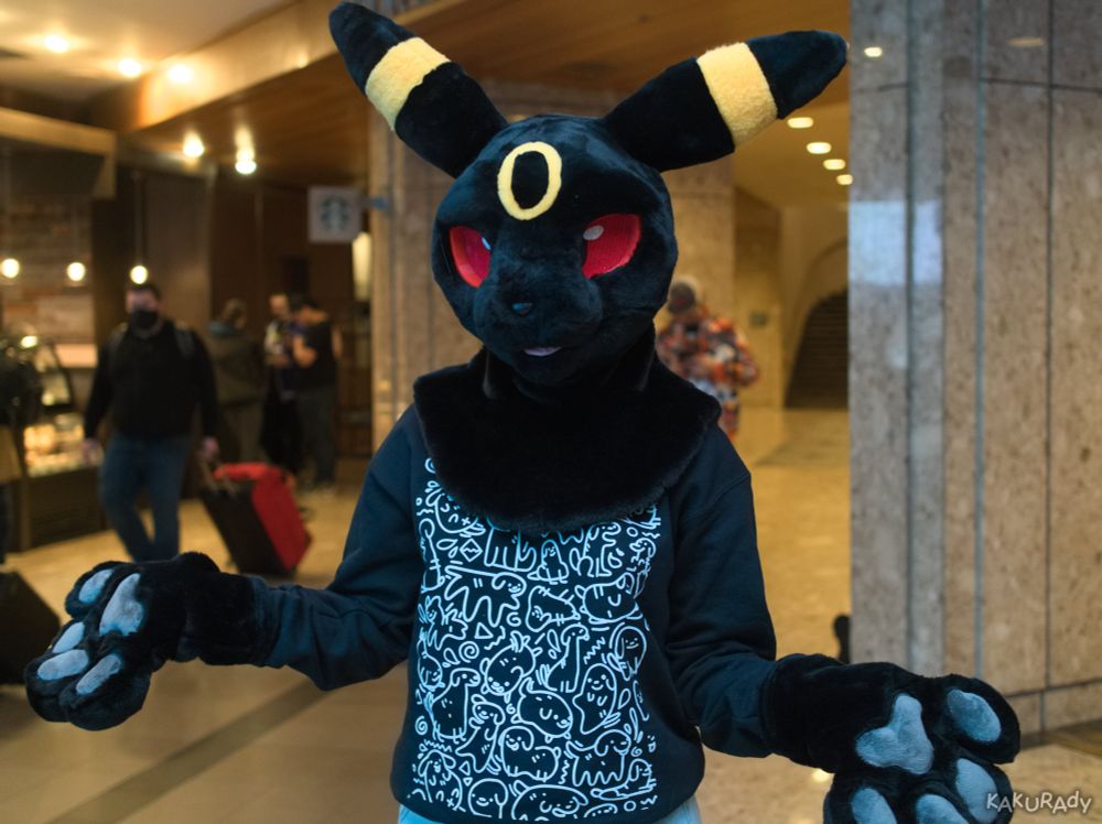 An Umberon fursuiter, wearing a black long sleeved shirt that has its front filled with white doodles of dogs, standing in a stone-clad hallway in front of a coffee shop booth, spreading their hands with elbows at the waist. 