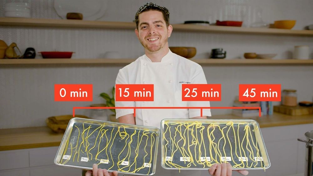 Chef Jack Logue stands in a kitchen holding two trays of pasta with timing labels showing 0, 15, 25, and 45 minutes to demonstrate different cooking stages.