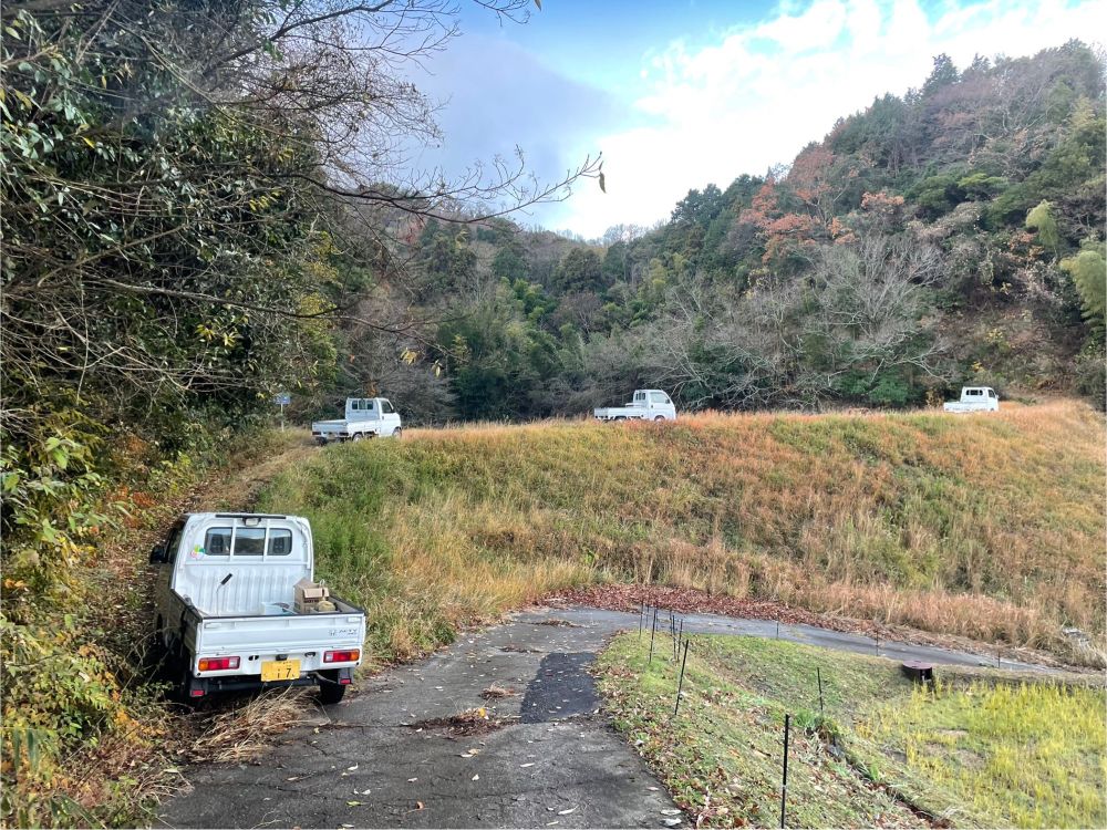 白い軽トラック4台が列をなしてため池の堤を走っている。背景は山林。