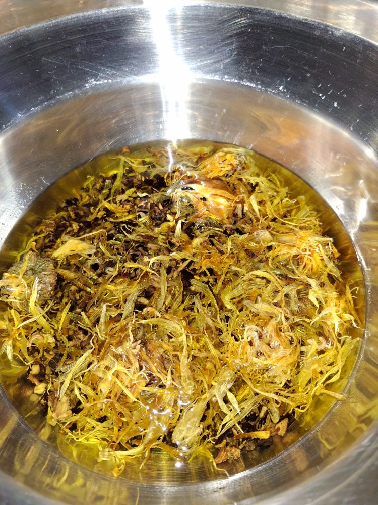 Stainless steel double boiler with various petals, leaves, and roots brewing in a melted ointment base. Looking very yellow.