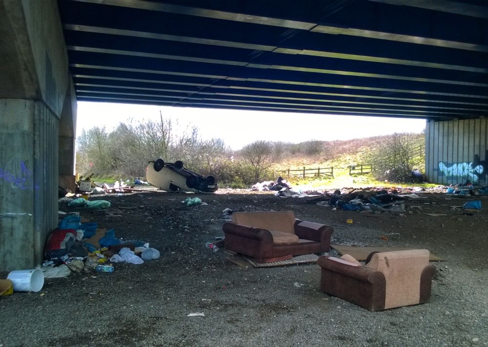 Below a motorway bridge, scrub land beyond. In the foreground two sofas facing each other. In the background a white van on its roof. Dumped rubbish all around.