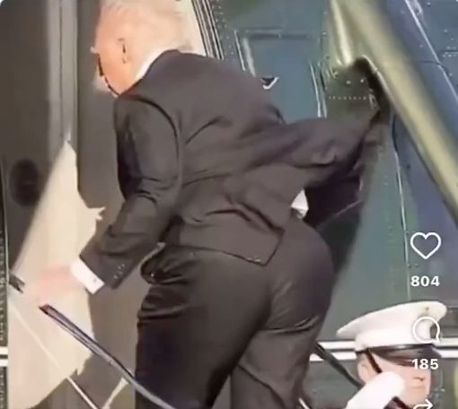 Um frame do vídeo abaixo que mostra trump com uma bunda gigante entrando num helicóptero tendo a bunda empurrada por um oficial.