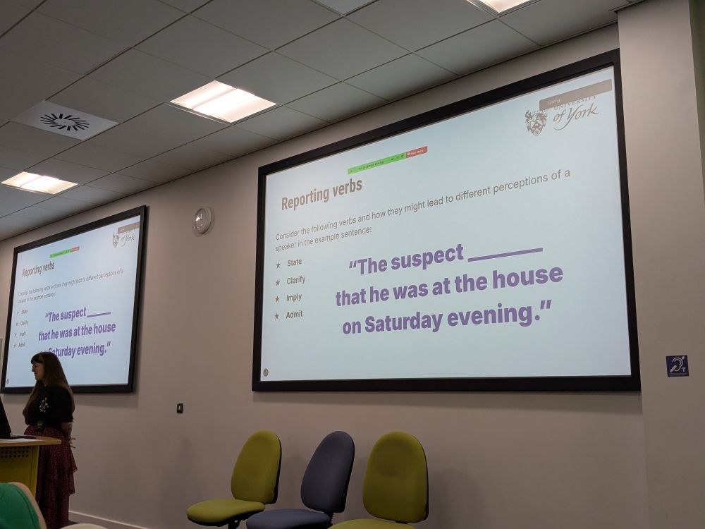 Lauren is standing at the front of the room under a large screen showing a slide with a task to reflect on how different reporting verbs can change the interpretation of reported speech