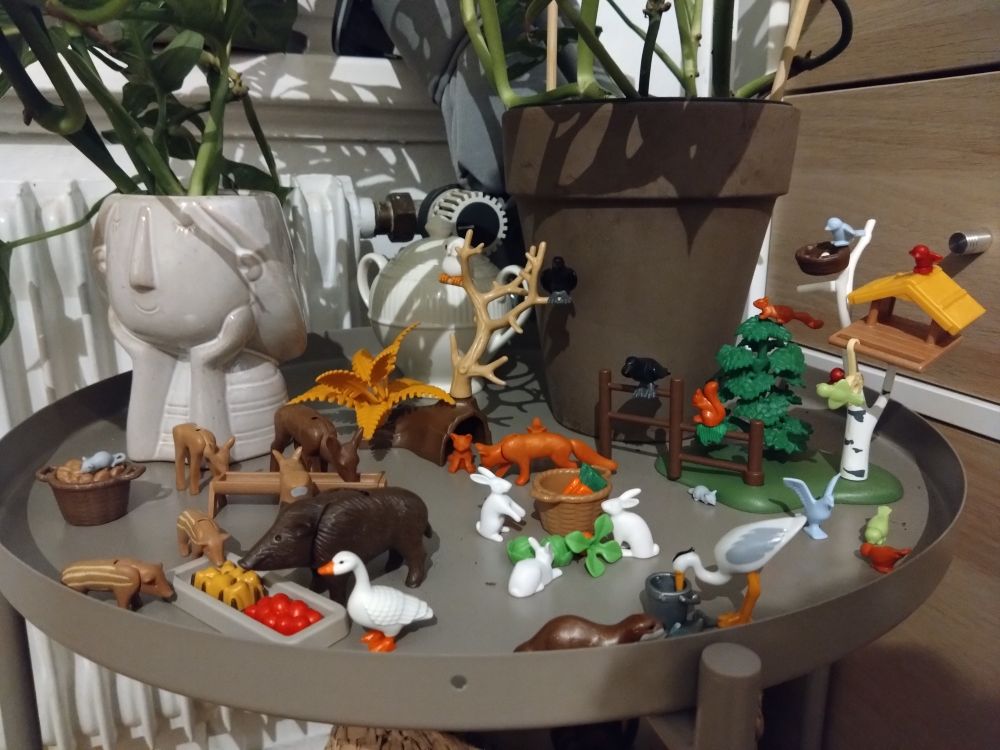 Auf einem Beistelltisch eine Szene aus Playmobil aufgebaut. Diverse Wildtiere, u.a. Rehe, Wildschweine, Füchse, Kaninchen, Mäuse, ein Otter, ein Kranich eine  Gans, an einer Art Futterstelle. In einem Baum bzw. Busch Krähen, eine Eule und Eichhörnchen, ausserdem ein Vogelfutterhaus mit einer Taube und mehreren kleinen Vögeln.