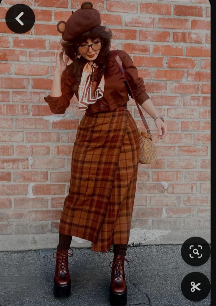 A woman in an outfit composed of a brown teddy bear hat, large rimmed glasses, a brown blouse, a long orange plaid skirt, beige purse and laced up elivated boots. 
