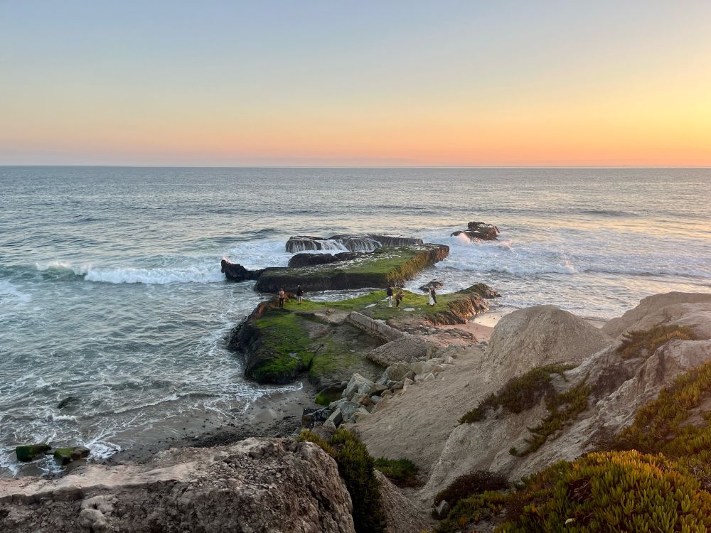 View of rocks descending into ocean from atop bluff, sunset visible to the side 