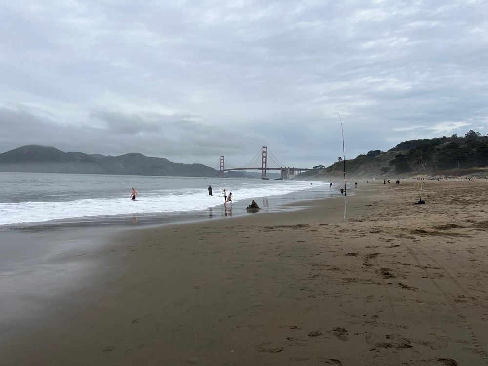View of Golden Gate Bridge from Baker Beach. Cloudy weather, ocean foam, and wet sand are visible.