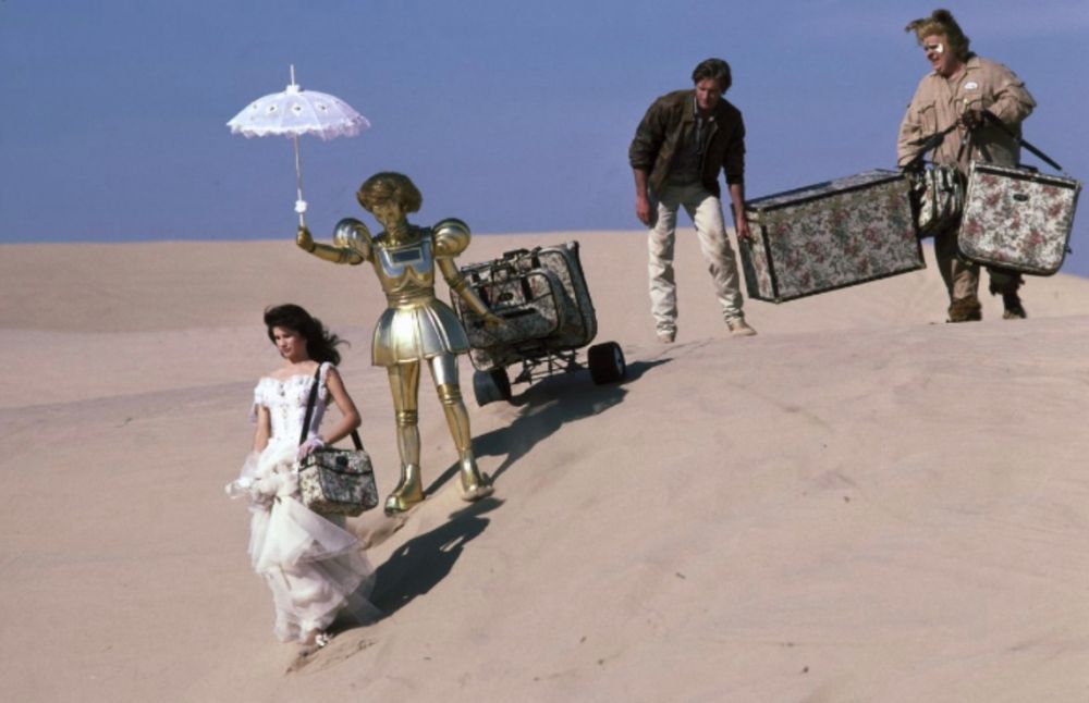 Szene aus „Spaceballs“: Prinzessin Leys im weißen Kleid wandert durch die Wüste, hinter ihr tragen ein Android und 2 Männer ihr Gepäck.
Der Android hält außerdem einen Sonnenschirm über die Prinzessin.