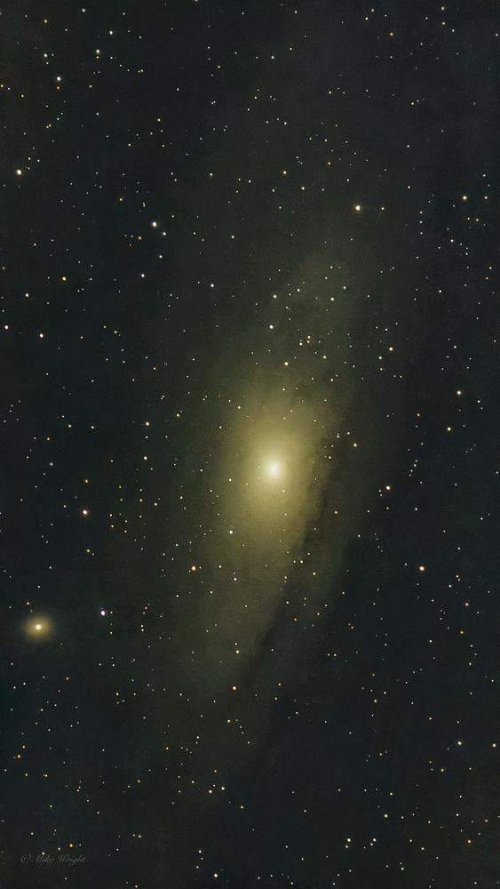 A deep-space photograph of the Andromeda Galaxy (M31), showing its bright, glowing core surrounded by a soft halo of dust and stars. The galaxy’s spiral structure is faintly visible, blending into the dark background filled with numerous distant stars. The image has a natural golden hue in the core, contrasting with the deep blackness of space.