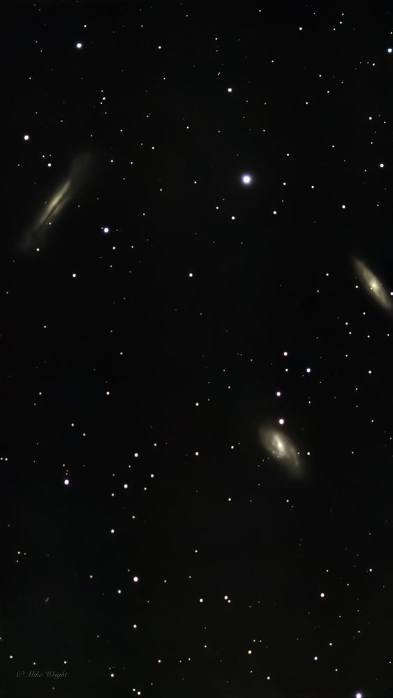 A deep-space photograph of the Leo Triplet, a group of three spiral galaxies—M65, M66, and NGC 3628—located in the constellation Leo. The galaxies appear at different angles, with NGC 3628 (left) showing an edge-on view, while M65 and M66 (right and bottom) display their spiral structures. The dark sky is scattered with numerous stars in the background.