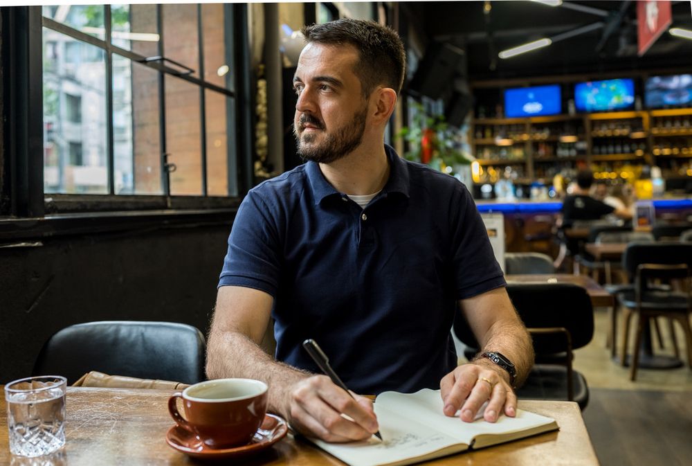 A man with short dark hair and a beard wearing a navy blue polo shirt sits thoughtfully at a wooden table in a café. He is writing in a notebook with a pen, with a brown coffee cup and glass of water beside him. The café interior features large windows, bookshelves, and blurred patrons in the background.