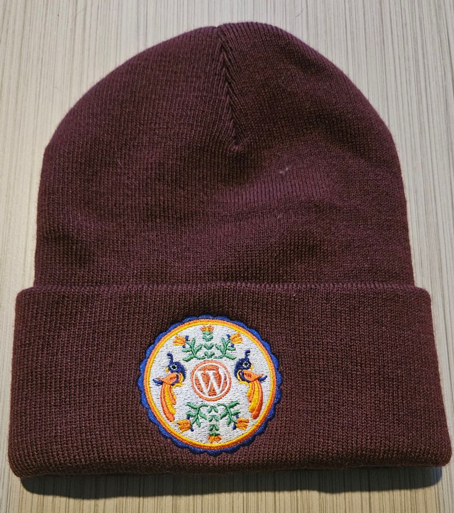 Maroon beanie. Circular patch on the front has an orange WordPress logo in the middle; two colorful, long-tailed birds, one to the left and one to the right; and stemmed flowers above and below with the stems toward the logo.