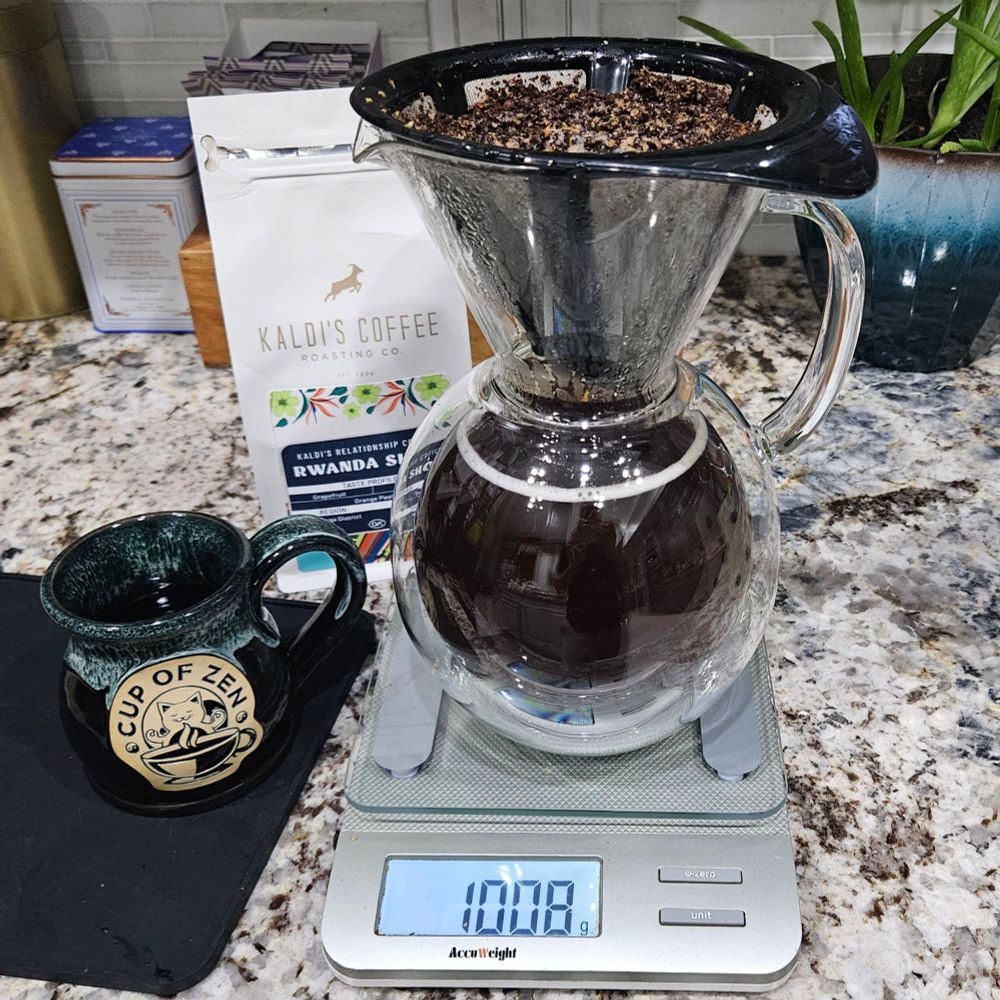 Rwanda Sholi whole bean coffee behind a coffee mug and a pourover carafe on a scale.