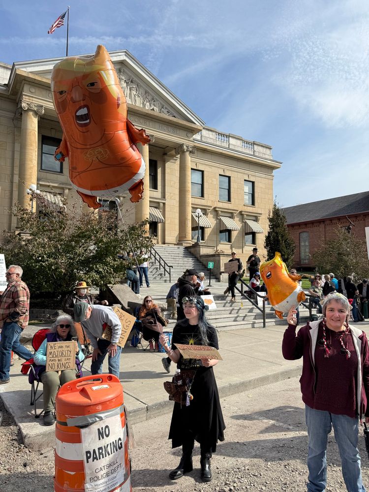 Woman in steampunk gear holding a mylar balloon of a baby Donald Trump in diapers in front of the Catskill, NY courthouse during the No Kings rally.