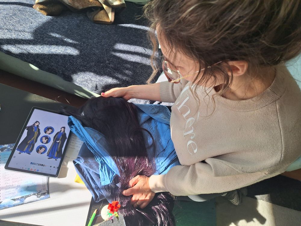 The picture shows a person from above in a beige sweater holding a dark purple wig against blue fabric and a tablet with a picture of the character Jinshi from Apothecary diaries 