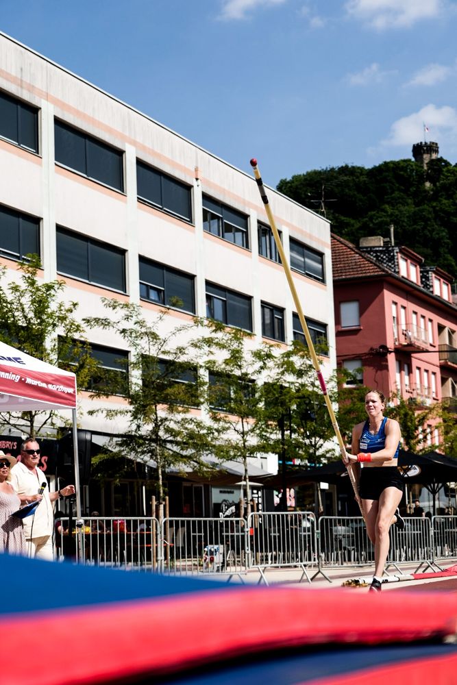 Jeune athlète féminine se lançant pour un saut à la perche sur la place d'une ville avec en arrière plan, la tour d'un château sur une colline 