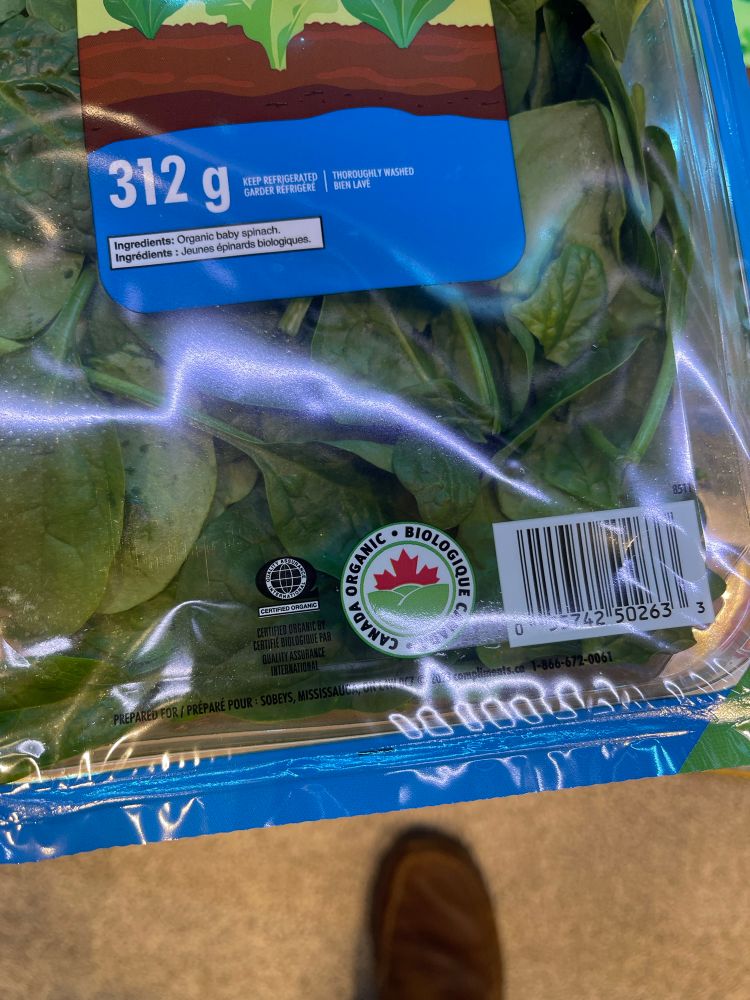 First of 3 photos of a package of Compliments spinach at Sobeys. On this package is a red and green symbol featuring a maple leaf in red and green rolling hills. The text in a circle around the symbols says Canada Organic. 