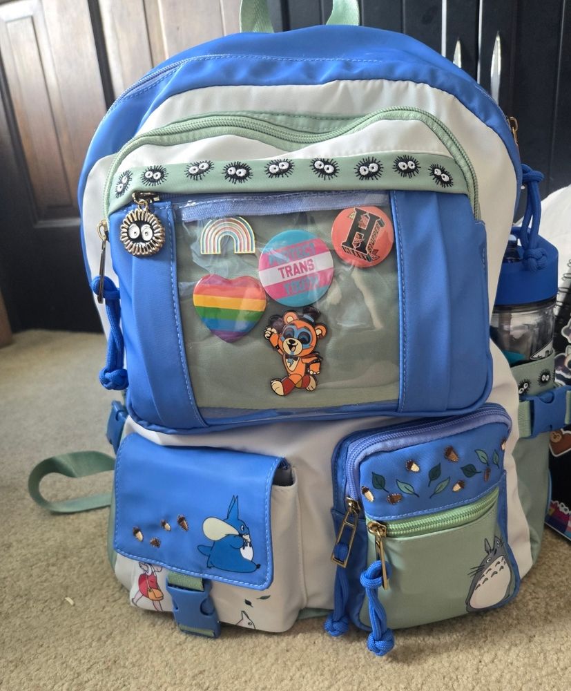 A multi pocket pastel green and light blue my neighbor totoro backpack, on the bottom left pocket is mei, with a white mini totoro, on the pouch, on the flap there is a blue mini totoro carrying a sack with acorns trailing behind it,  on the right pocket is totoro himself on the bottom with various leaves and acorns on the top. it has a plastic sleeve in the upper middle where a transflag of rainbow pin, "protect trans youth" pin,  a "H" for Heartstopper pin, a rainbow heart pin,  and a glamrock freddy pin sits,  one of the zippers is a soot sprite, and the top of the plastic lining is fabric with soot sprites on it. it has two drink pockets on the side with clasps to hold my bottle snuggly 