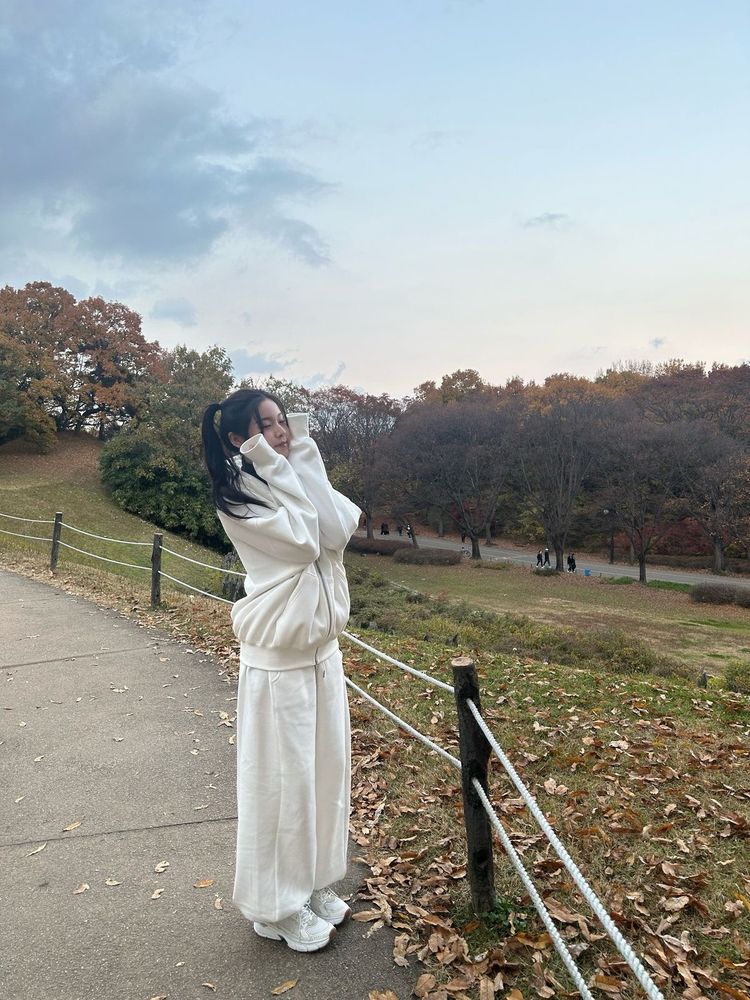 Photo of kyujin from kpop group nmixx in a white sweatsuit at a park framing her face with her sweater paws