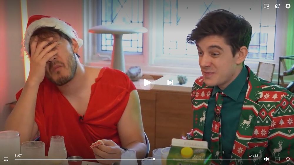 A screenshot of the Unus Annus video "Santa's Mukbang (Drinking 1 Gallon of Eggnog)". Markiplier is holding a sugar cookie in one hand and has his other on his face. He's wearing a Santa hat, and a large Santa gift bag around his torso that he used a knife to cut holes into. 
Ethan is to the right of him with a pained expression on his face. He's wearing a green Christmas suit with red stripes and white designs like reindeer, snowflakes, and trees. 
They're both sitting at a table with a lot of empty eggnog glasses surrounding them. 