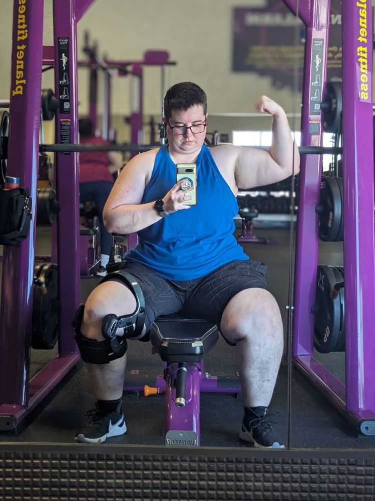 Picture of Jack Morgan Lavandowska sitting on a workout bench at a Smith machine, wearing a blue tank top, black shorts, and a knee brace. They are flexing their right bicep while looking at their phone camera. 